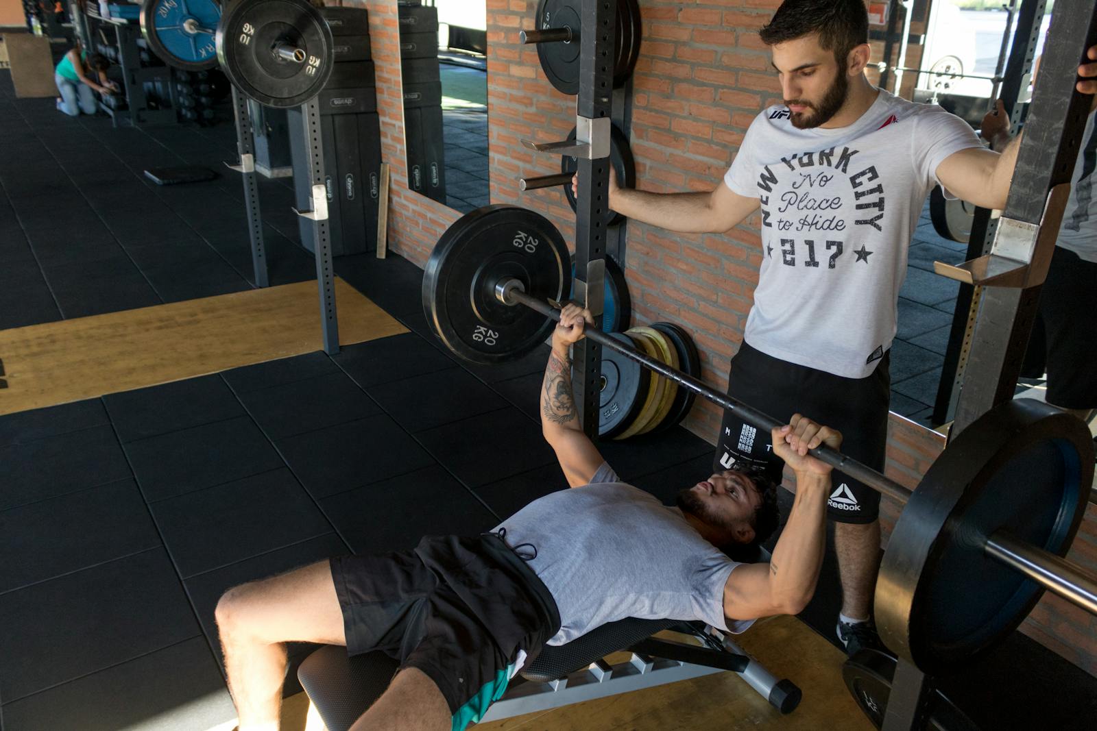 A strong man lifts weights on a bench while a trainer assists in a modern gym setup.